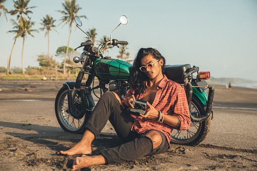 Man sitting near motorcycle at the beach while using instant camera