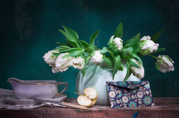 Classic still life with white tulips, half an apple and an envelope on old wooden table on green background