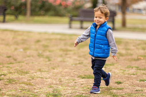 Cute boy in a blue vest running in the park holding out his arms like an airplane. Selective focus.