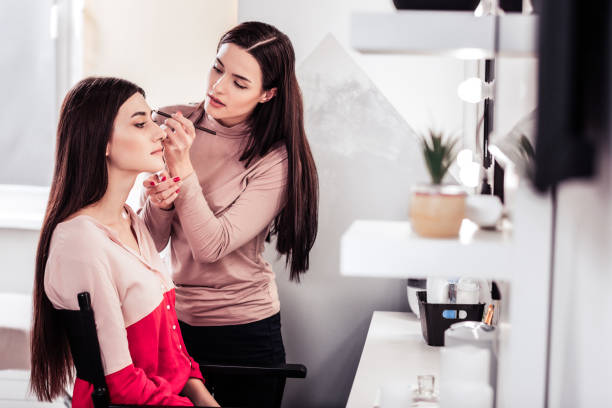 Skilled talented makeup artist focusing on her work Real talent. Skilled talented makeup artist standing near her client while focusing on her work septum stock pictures, royalty-free photos & images