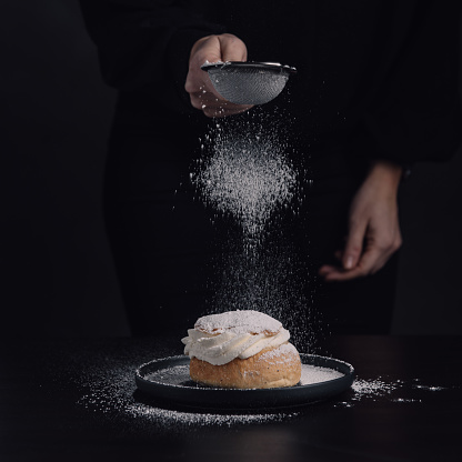 Swedish semla typical wheat bun with cream\nPhoto taken in studio, woman spreading powder sugar on fresh semla\nStudio shot, dark low key photo