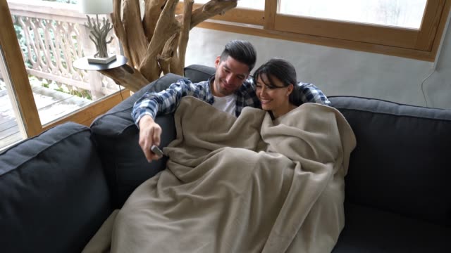 Loving young couple at a winter lodge lying down on couch under a blanket watching tv talking