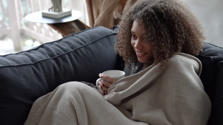 African american woman watching tv covering herself with a blanket and drinking coffee in a cold winter morning very happy