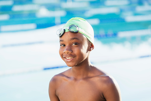 A 9 year old African-American boy at a swimming pool wearing a swim cap, smiling. The pool is set up for competition with lane ropes.