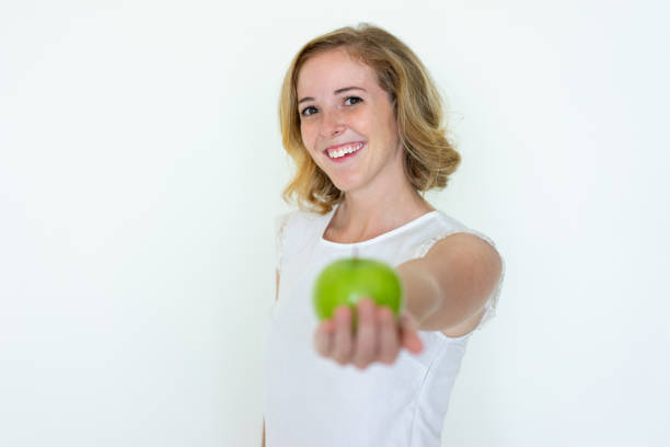 Smiling young pretty woman offering blurred green apple. Lady looking at camera. Raw food concept. Isolated front view on white background.