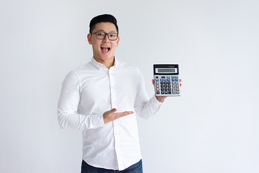 Excited Asian man holding and showing calculator. Young guy standing and looking at camera. Finance concept. Isolated front view on white background.