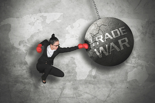 Image of young businesswoman wearing boxing gloves while jumping and hitting a pendulum with trade war text