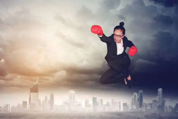 Picture of young female entrepreneur wearing boxing gloves while jumping and punching through at outdoor