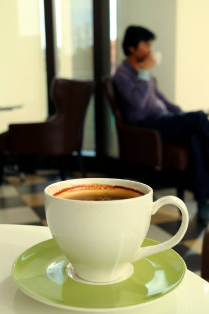 A Cup of Hot Coffee on a White Table with Blurry Drinking Coffee Man in Background, Relaxing time