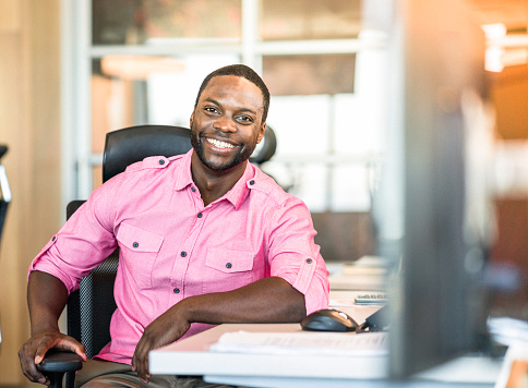 Portrait of smiling male professional sitting on chair at desk. Confident young CEO of a small computer programming businesss in office. He is wearing pink shirt.