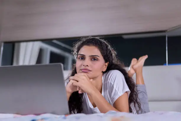 Picture of curly hair woman looking at the camera while lying with a laptop on the bed. Shot at home