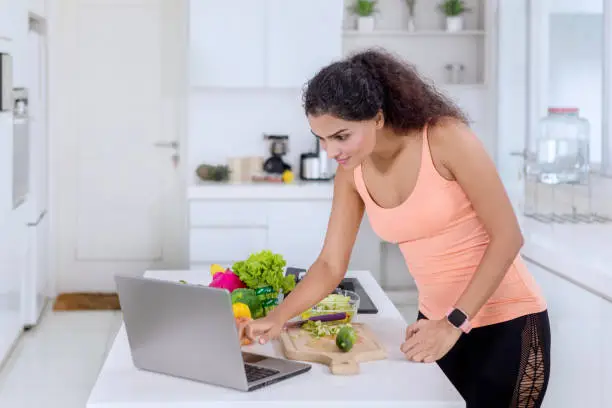 Side view of a pretty girl using a laptop while preparing salad in the kitchen