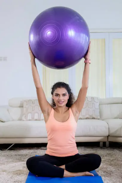 Picture of curly hair woman lifting a yoga ball while exercising in the living room. Shot at home