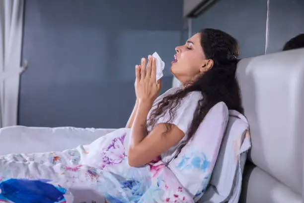 Portrait of curly hair woman sneezing with tissues in her hands while sitting on the bed