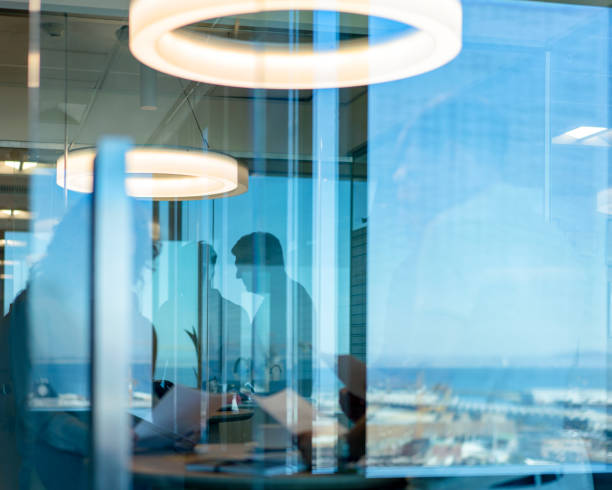 Business colleagues discussing over documents in board room. Professionals are seen through window. They are standing at workplace.