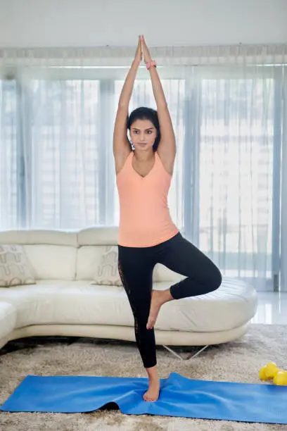 Full length of curly hair woman wearing sportswear while exercising yoga with tree pose at home