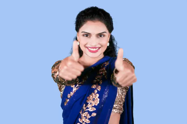 Pretty Indian girl smiling at the camera while showing thumbs up. Shot in the studio with blue background