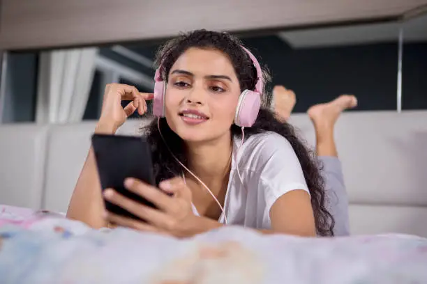 Image of pretty girl is hearing music by using headset and smartphone while lying on the bed