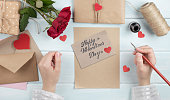 Valentines day theme. Top view of female hands writes greetings. Packed gifts, roses  and envelopes on shabby wooden table
