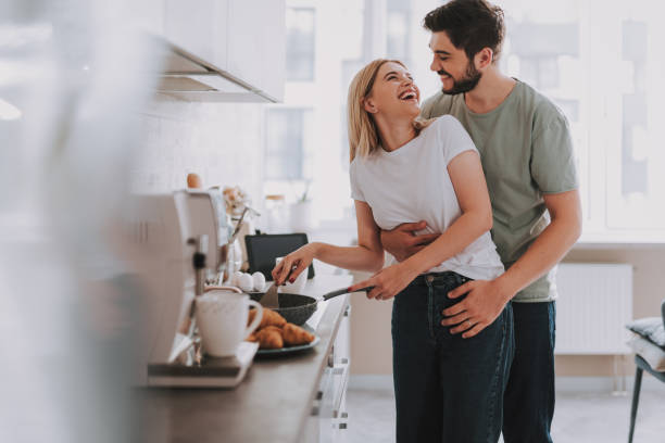 Young cheerful spouses enjoying morning time together Cooked with love. Cute laughing blonde frying something in skillet. Her bearded husband is hugging her from behind wife stock pictures, royalty-free photos & images