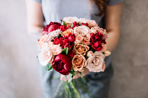 Very nice young woman holding a beautiful flower wedding bouquet of fresh roses and  peonies, in pastel cream and vivid red colors on the grey background