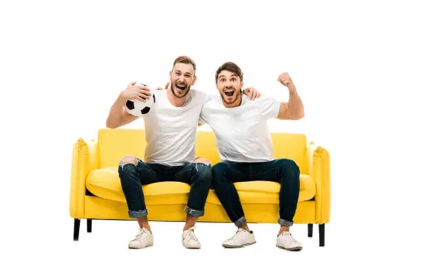 Photo of excited young man sitting on couch with soccer ball isolated on white