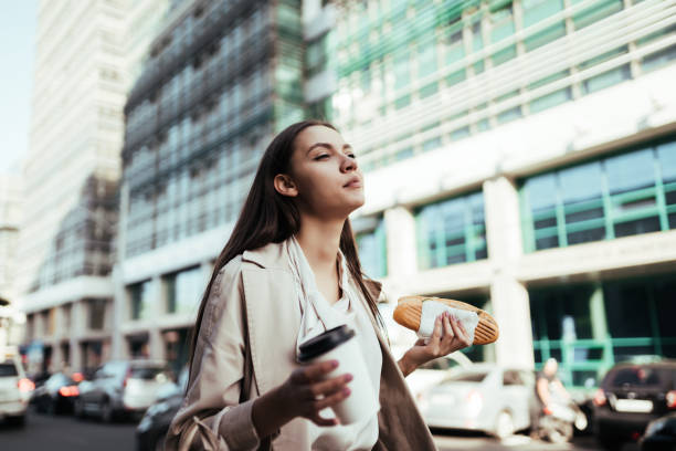 joyful girl walks down the street and snacks on a sandwich against the background of office buildings - food currency breakfast business imagens e fotografias de stock