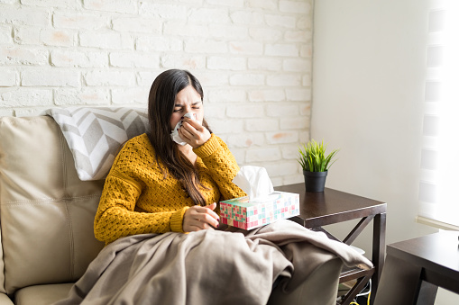 Sick woman blowing her nose in tissue while sitting on sofa at home