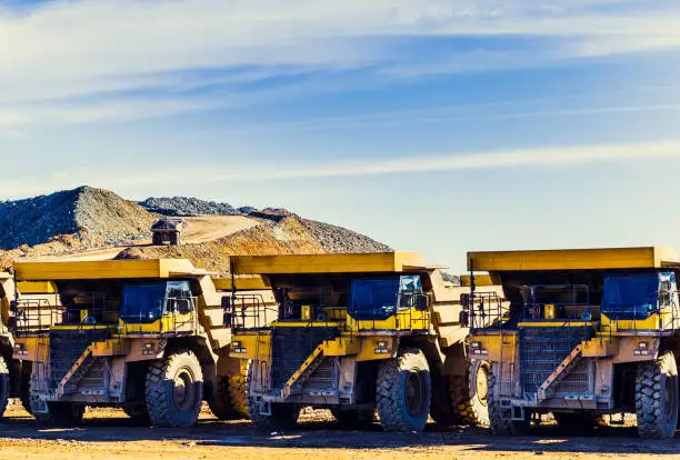 Big yellow dumper trucks placed in a row in the mine with dumper trucks laden with ore on the quarry road with blue sky and clouds in the background