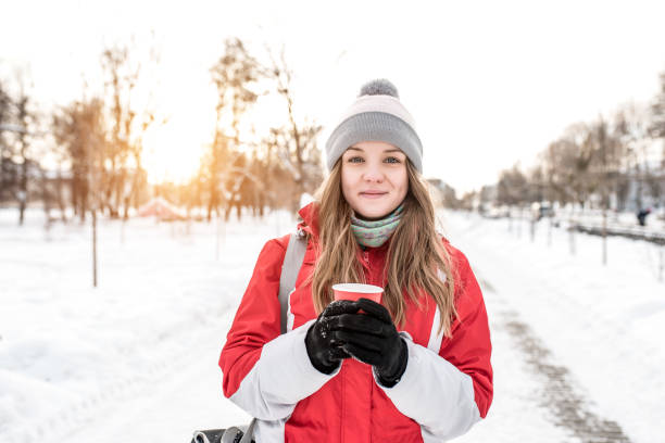 a garota no inverno no gatilho vermelho e cabelo longo. em suas mãos segurando uma xícara de café ou chá. no inverno na cidade sobre um fundo de um pôr do sol ou nascer do sol. sorrisos felizes. espaço livre para texto. - power walking flash - fotografias e filmes do acervo