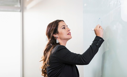 A confident Hispanic businesswoman standing in an office board room, looking up as she writes on a whiteboard, giving a presentation. She is a mid adult woman in her 30s, well-dressed in businesswear.