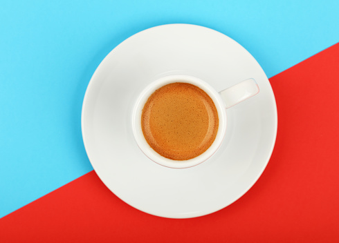 Close up one white cup full of espresso coffee on saucer over colorful red and blue background, elevated top view, directly above