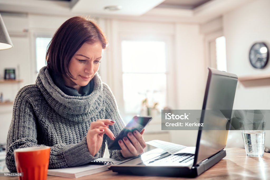 Woman typing text message on a smart phone Woman wearing grey sweater typing text message on a smart phone while sitting by the table in the office Computer Stock Photo