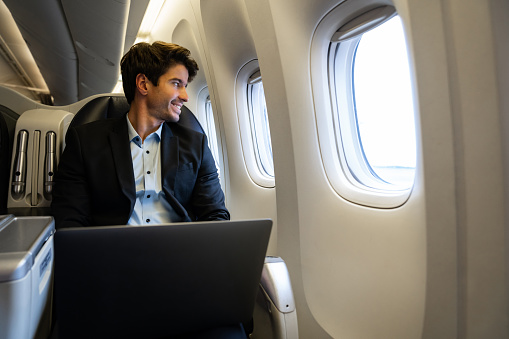 Portrait of a business man traveling by plane and working on his laptop computer while looking through the window smiling - business trip concepts