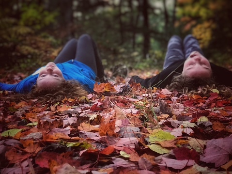 Two Caucasian, cute girlfriends or sisters, laying in autumn leaves, on a trail, in a dense forest. One girl is looking back towards the camera, the other has eyes closed making a silly face. Selective focus and soft blurs added with digital enhancements. Photo taken in Northern Wisconsin.