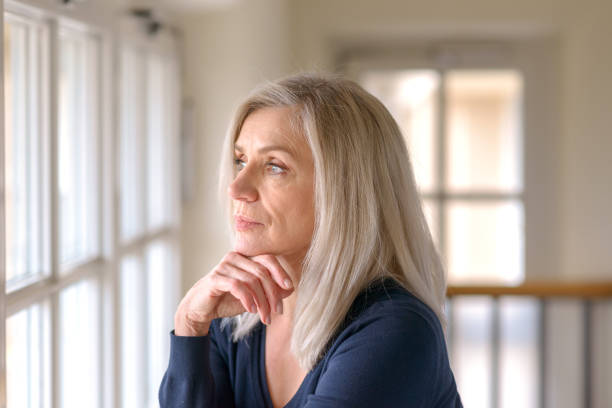 Pretty thoughtful woman with serious expression Attractive thoughtful woman with serious expression standing with her hand to her chin staring quietly out of a large window staring stock pictures, royalty-free photos & images
