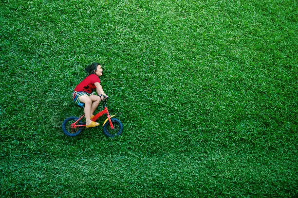 Rear View of a Happy Asian Kids. Girl on Bicycle Lay Down at Green lawn in Summer Day. Excited Asian Children in her Outdoor Activity