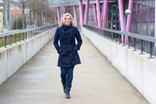 Trendy blond woman in a stylish blue outfit walking along a walkway in town approaching the camera with a friendly smile