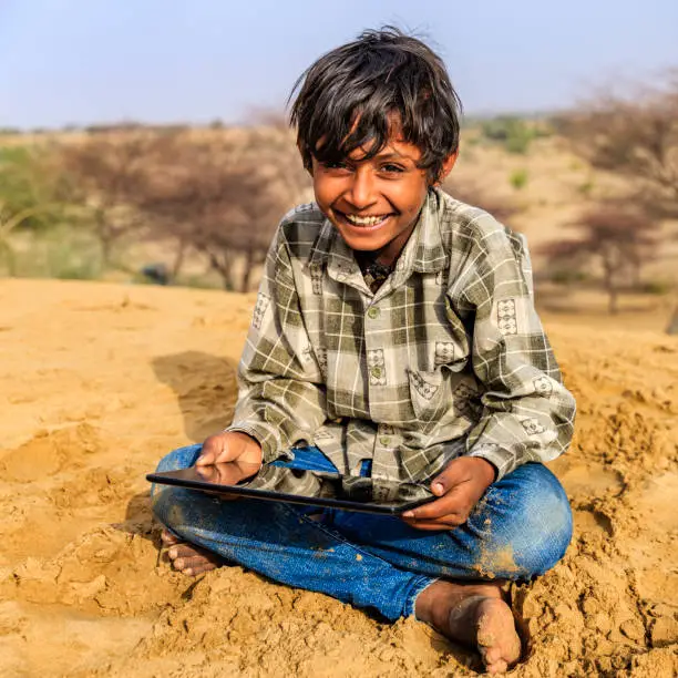 Happy young Indian boy sitting on a sand dune and using digital tablet in desert village, Thar Desert, Rajasthan, India.