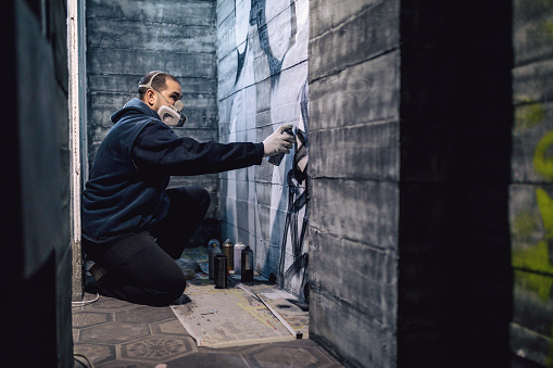 Artistic graffiti artist using a spray paint on a wall indoors, making a street art graffiti.