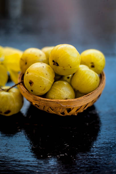 Close up of organic raw Indian gooseberry or amla or Ribes uva-crispa in a vegetable and fruit basket made up of bamboo on wooden surface with some leaves of Rangoon's creeper. stock photo