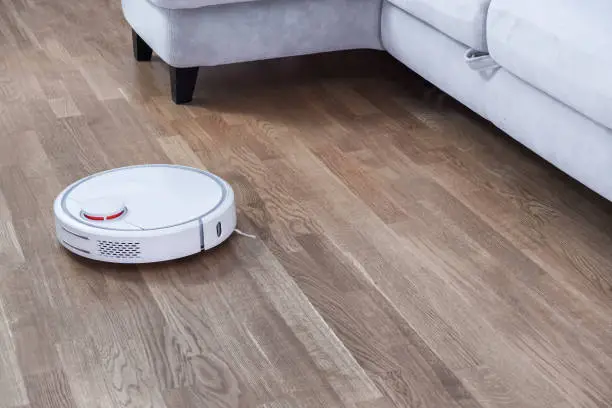 Robotic vacuum cleaner runs near sofa in room on laminate floor. Robot controlled by voice commands to direct cleaning. Modern smart cleaning technology housekeeping.
