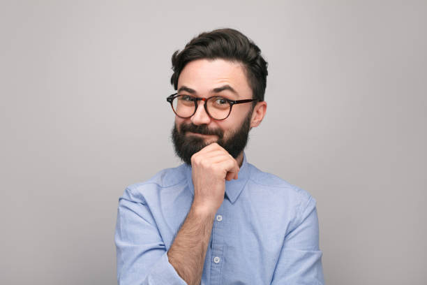 Playful bearded guy in eyeglasses Stylish casual man with beard wearing eyeglasses and looking skeptically at camera on gray background. suspicion stock pictures, royalty-free photos & images