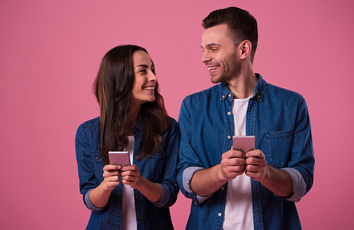 Cheerful young couple standing isolated over pink background, holding smart phones in casual wear