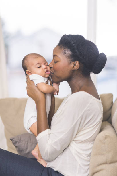 Beautiful African American mother holds newborn baby in the living room A beautiful young African American mother gently holds her infant daughter up in the air with both hands and kisses her cheek. The baby's eyes are wide open and she looks happy. They are sitting on a couch in their living room. biracial newborn stock pictures, royalty-free photos & images