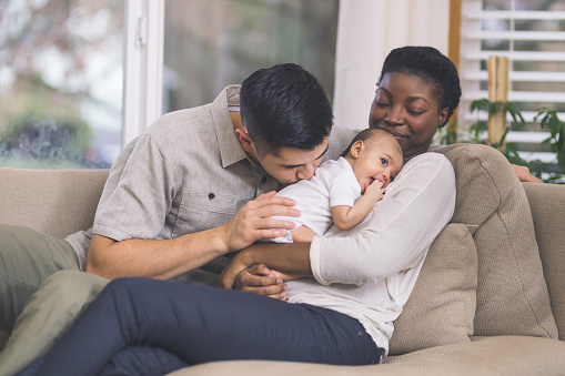A beautiful young African American mother gently holds her infant girl to her chest. The baby has her hand by her mouth and her eyes are wide open. Dad has his arm around mom and is looking at her affectionately.