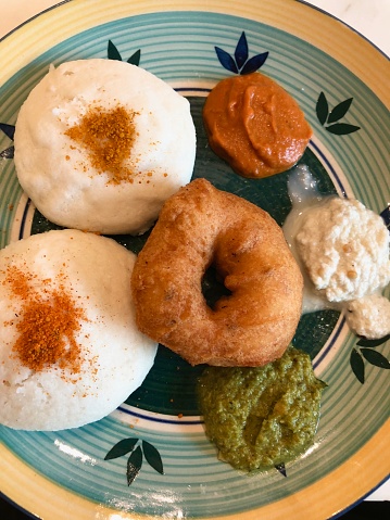 This photo includes traditional South-Indian breakfast in hotel restaurant buffet in Delhi, India. Which consist of idli, medu vada donut / doughnut with ginger tamarind chutney, coconut and coriander chutneys in a dish. Also Idli is sprinkled with gun power masala .