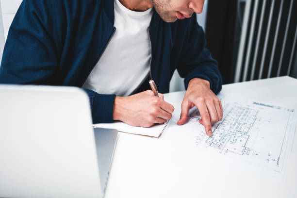 istock High view of hand hold notepad of businessman while working at office and using notebook. Project business concept. Young designer sitting in office and working with blueprint plan 1096808830