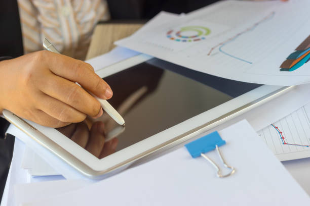 istock Hand of business analyst holding pen is analyzing graphs about marketing on tablet. Unfinished paperwork stacked in archive with color binder paper clips. Business and Education concept. 1096812656