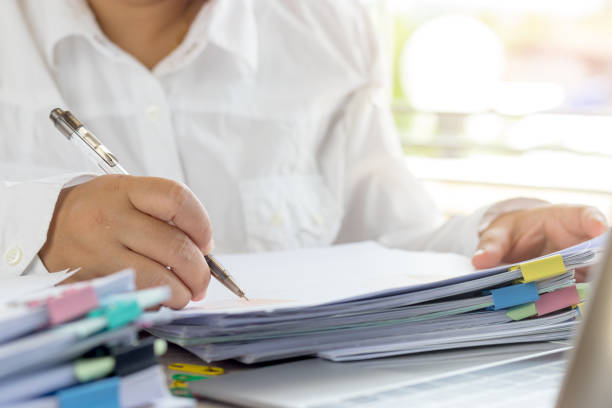 istock Teacher holding pen by hand for checking student homework assignments on desk in school office. Unfinished paperwork stacked in archive with color binder paper clips. Education and business concept. 1096812974
